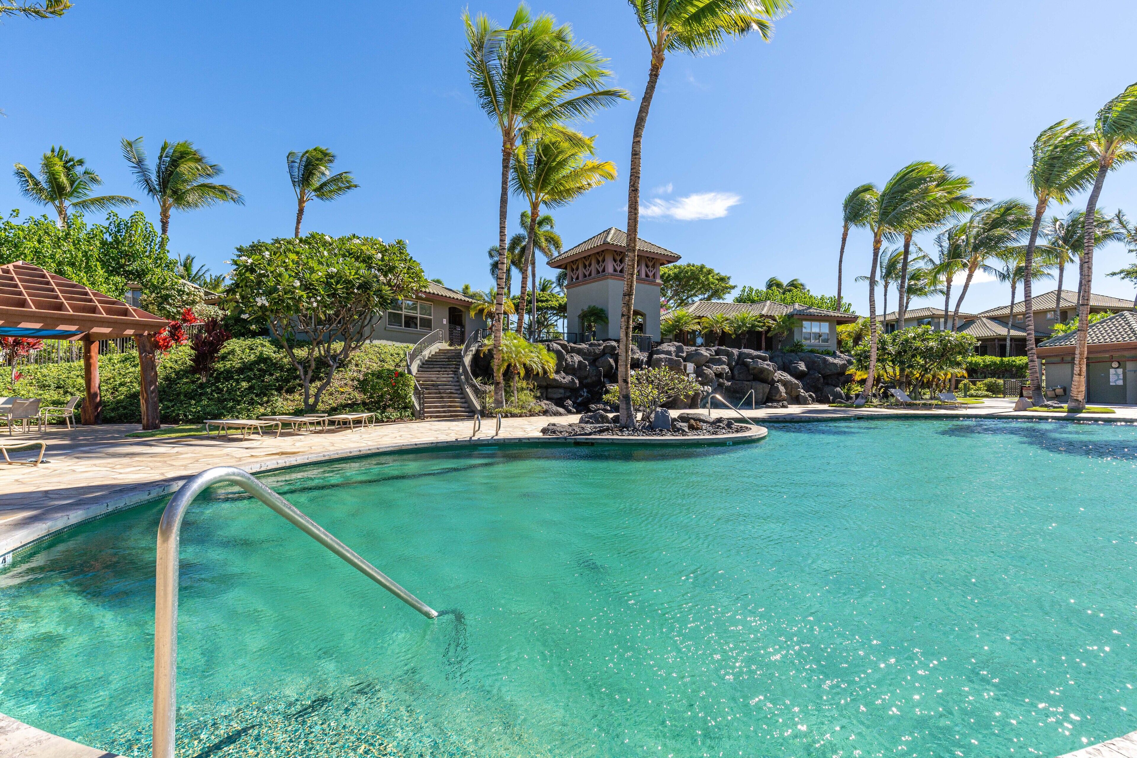A clear swimming pool with a metal ladder, surrounded by palm trees and a building with a red roof.