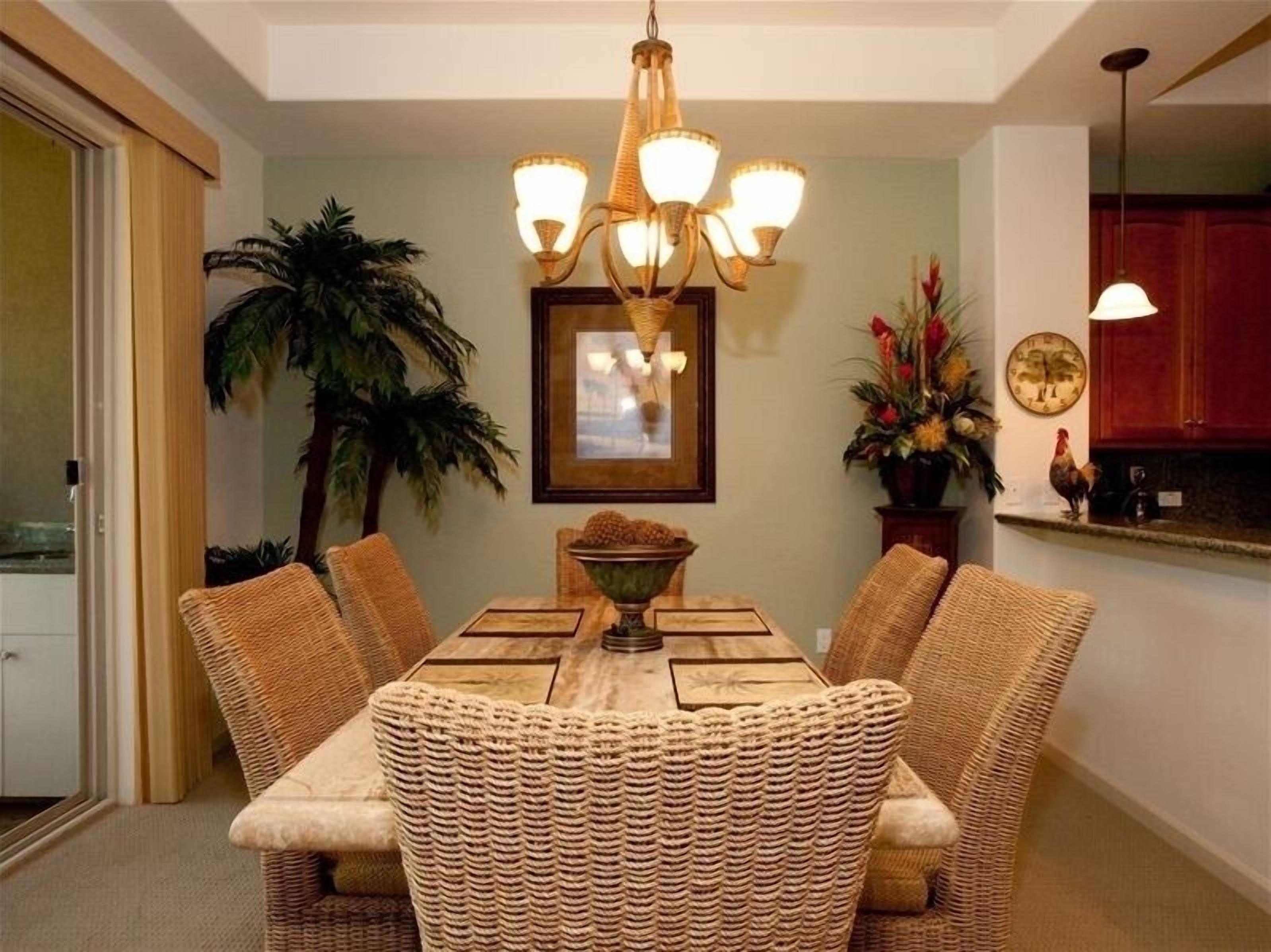 Un coin repas avec une table et des chaises en osier, un lustre, un tableau encadré et une plante en pot.