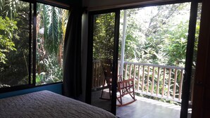 A bedroom with a large window, a bed, and a wooden rocking chair on a balcony with a view of dense foliage.