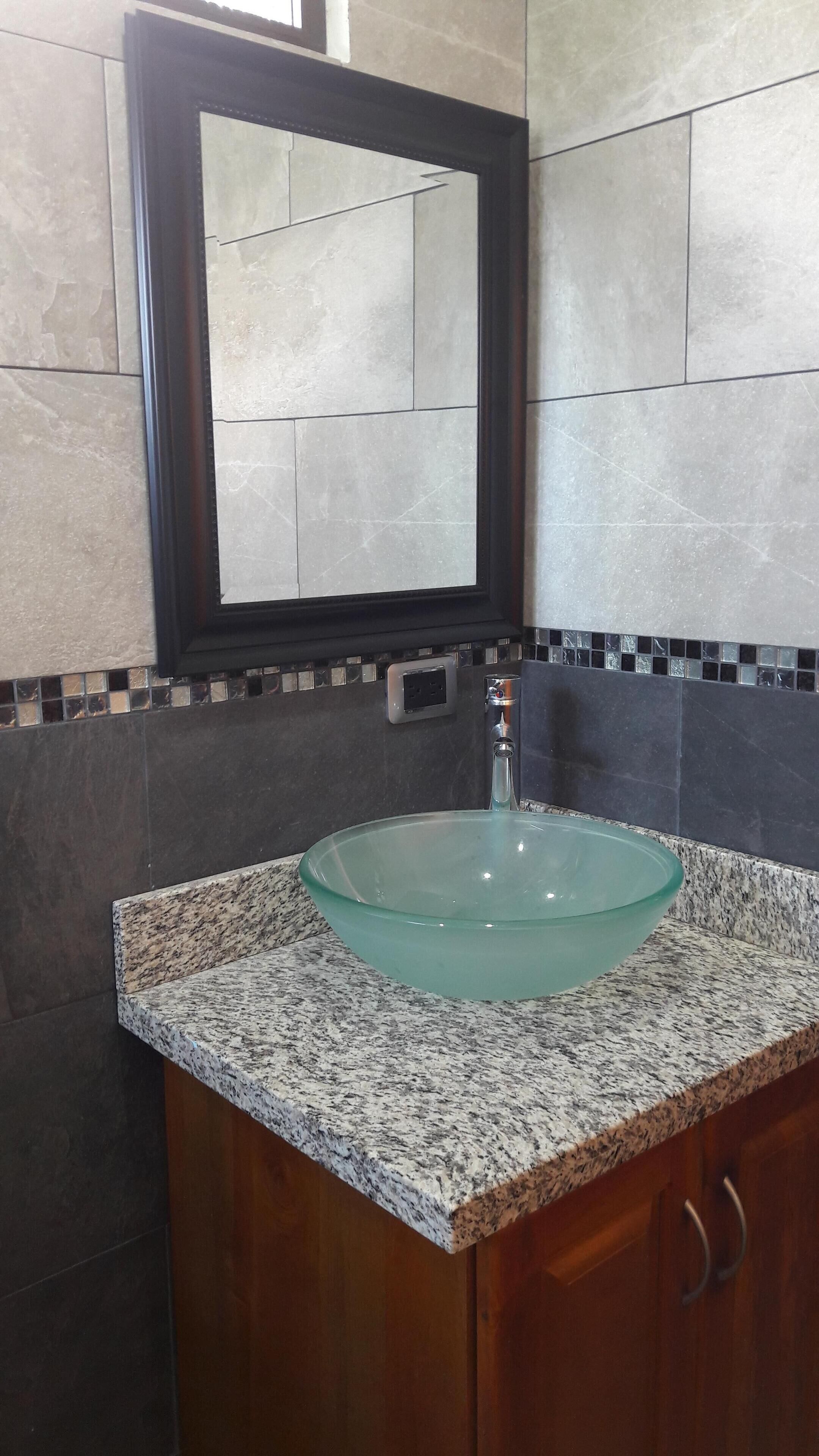 A bathroom with a glass sink, granite countertop, dark tiles, and a mirror.