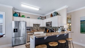 A modern kitchen with white cabinets, a stainless steel refrigerator, a microwave, and a coffee maker. There are three wooden stools and a patterned rug. - Orange Beach Luxury Corner Condo (Orange Beach)