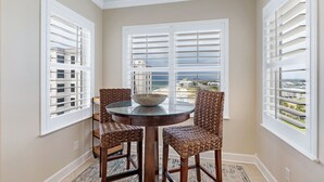 A dining area with a round table and four wicker chairs, large windows with white shutters, and a view of the ocean and buildings. - Orange Beach Luxury Corner Condo (Orange Beach)