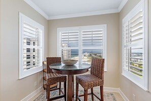 Un coin repas avec une table ronde et quatre chaises en osier, de grandes fenêtres avec des volets blancs, et une vue sur l’océan et les bâtiments.