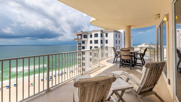 A balcony with a view of the beach and ocean, featuring a table and chairs, and a building in the background. - Orange Beach Luxury Corner Condo (Orange Beach)