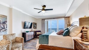 A bedroom with a bed, a ceiling fan, a TV, a dresser, and a view of the ocean. - Orange Beach Luxury Corner Condo (Orange Beach)