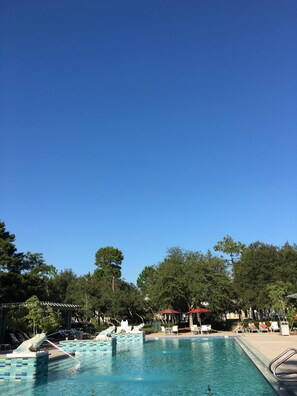 Une piscine dont les bords sont recouverts de carrelage, avec des chaises longues et un kiosque, le tout situé dans un cadre verdoyant qui ressemble à un parc.