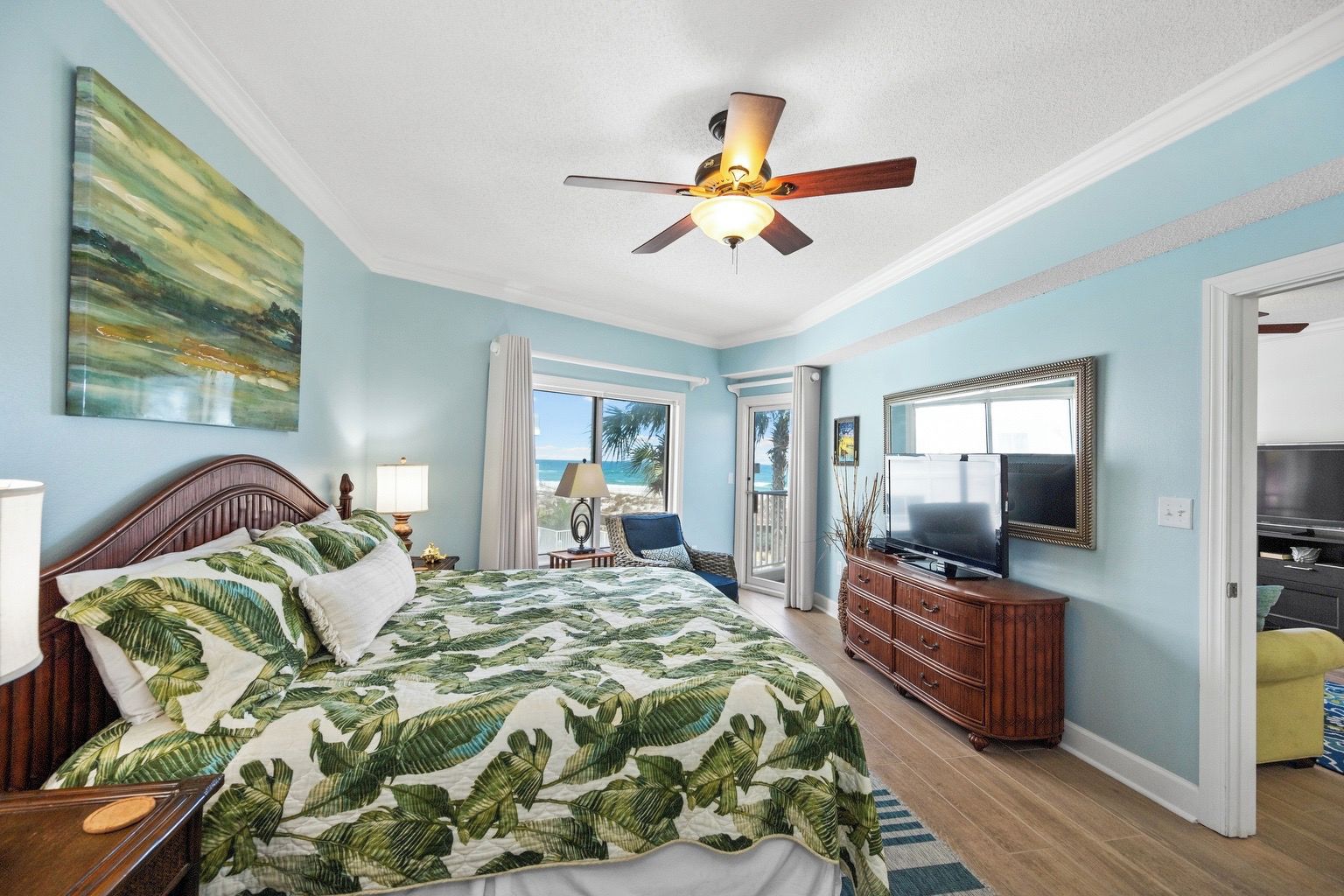 A bedroom with a bed, a ceiling fan, a TV, and a view of the beach.