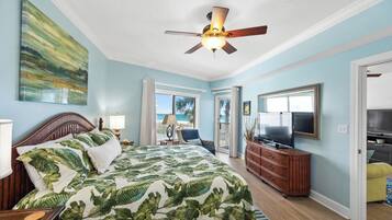 A bedroom with a bed, a ceiling fan, a TV, and a view of the beach.