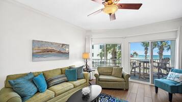A living room with a green sofa, a blue armchair, and a ceiling fan. There is a painting on the wall and a view of the ocean through the glass doors.
