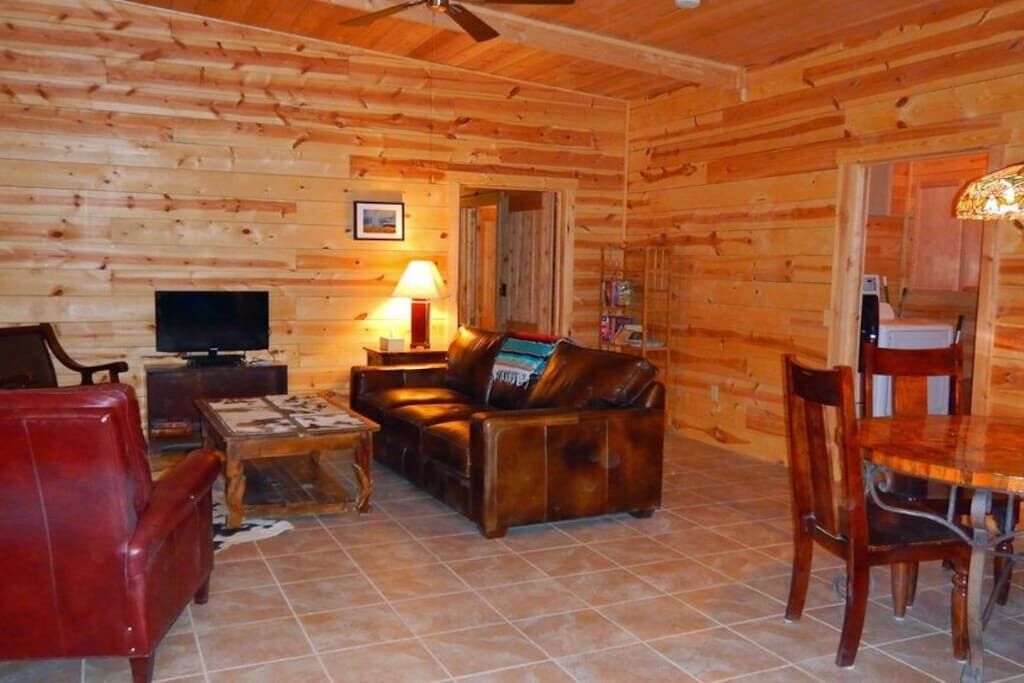 L’intérieur d’un chalet en bois, avec un canapé en cuir, un téléviseur, une table à manger et des chaises, ainsi qu’un ventilateur de plafond.