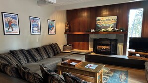 Een woonkamer met een hoekbank, een open haard, ingelijste posters en een televisie.