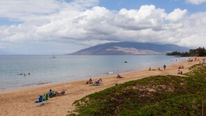 Una playa con gente tomando el sol y nadando, un velero en el agua y una montaña al fondo.