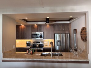 A modern kitchen with dark wood cabinets, stainless steel appliances, and a marble countertop. - Luxury 3BR Ocean View, 2 Lanai's, Steps to Kamaole I Beach! Hotel Zoned. (Kihei)