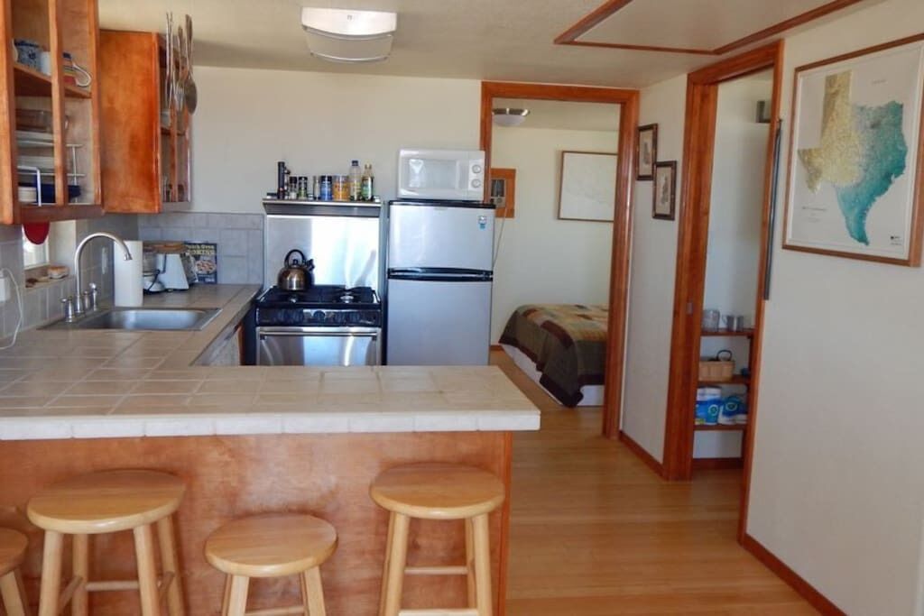 Cocina compacta con estufa, lavamanos y refrigerador, taburetes de madera en la barra, y un pasillo que conduce a un dormitorio.