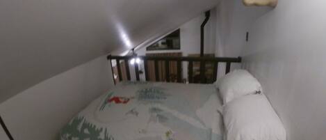 Een kleine, schemerige zolderkamer met een schuine wand, een bed met een beddegoed met dessin, een houten hoofdbord en een op de wand bevestigde hertenkop.