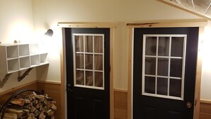 Une pièce avec deux portes noires, une pile de bois de chauffage et une étagère fixée au mur, sur laquelle sont posés divers objets.
