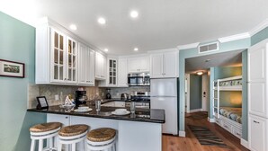 A kitchen with white cabinets, a central island with stools, stainless steel appliances, and a wooden floor. - AMAZING- beach condo overlooking Pool & Gulf of Mexico 2309 (Galveston)