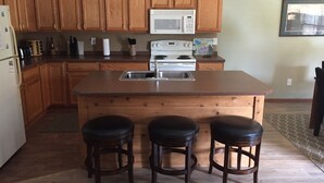 A kitchen with wooden cabinets, a white microwave, a stove, and a sink. There are three stools in front of the island.