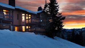 Un chalet à plusieurs étages doté de balcons, entouré d’un paysage enneigé et illuminé par un ciel au coucher du soleil.