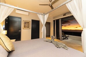 Une chambre à coucher avec un lit, une table de chevet, une lampe, une fenêtre avec des rideaux et une vue sur le coucher de soleil.