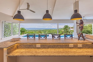 A modern kitchen with granite countertops, pendant lighting, and a view of a pool and greenery. - Luxury 6BR Ocean-View Villa  Infinity Pool, Spa, Concierge & Full A/C (Uvita)