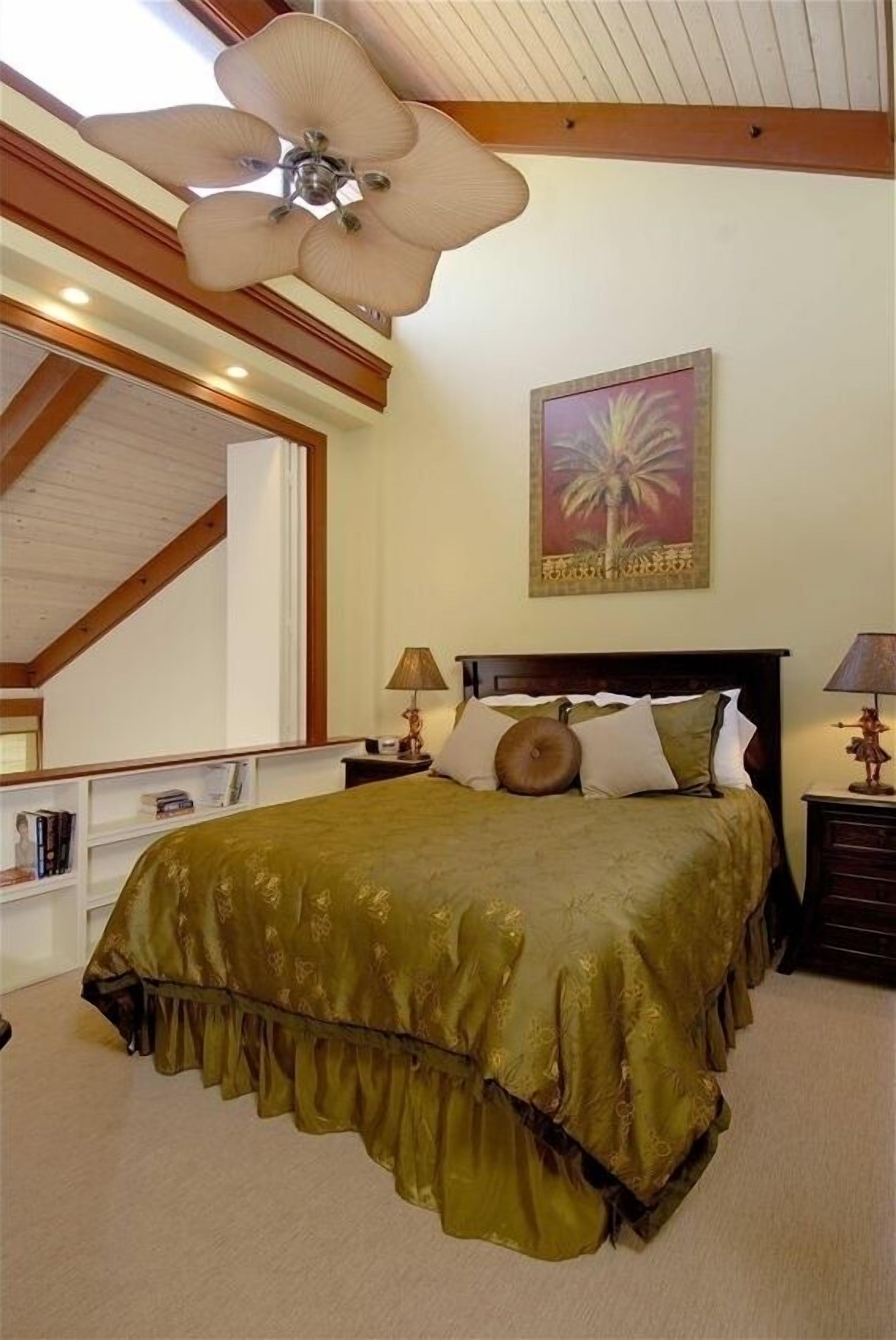 A bedroom with a large bed, a ceiling fan, a painting of a palm tree, and a bookshelf.