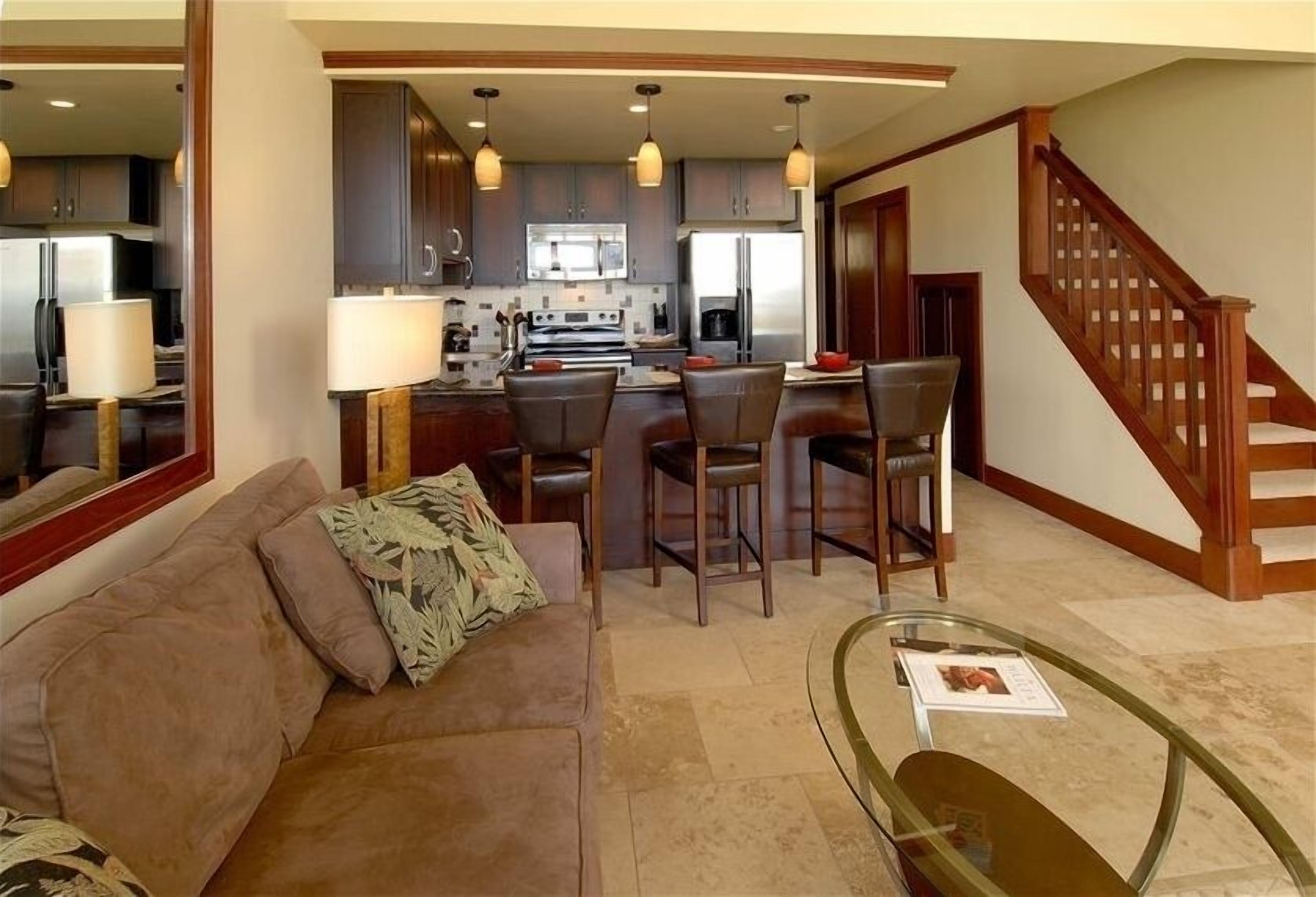 A spacious living room with a brown sofa, a glass coffee table, and a kitchen area with dark cabinets and modern appliances.