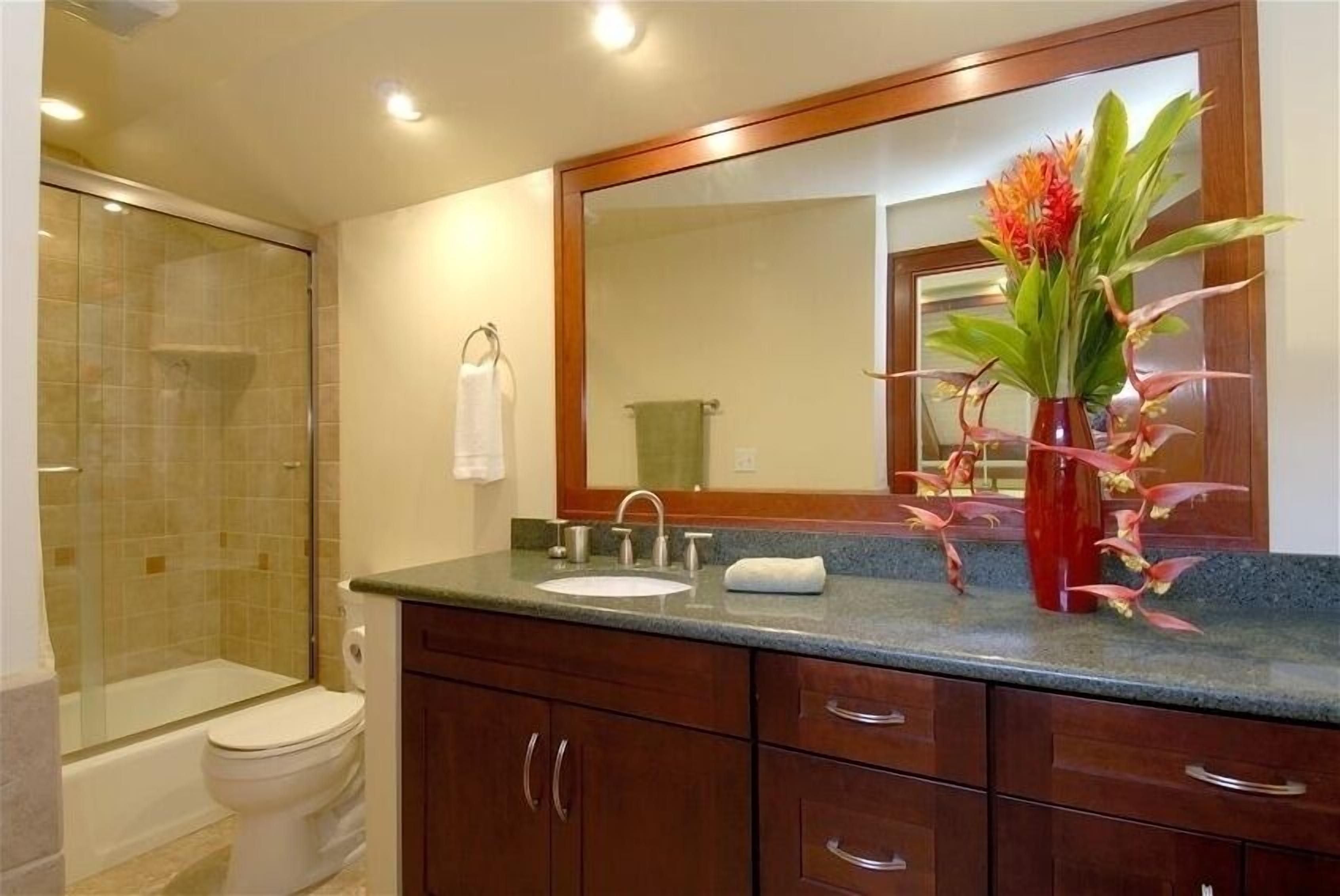 Une salle de bain avec une douche à parois de verre, des toilettes, un lavabo avec un miroir au-dessus et un vase avec des fleurs posé sur le comptoir.