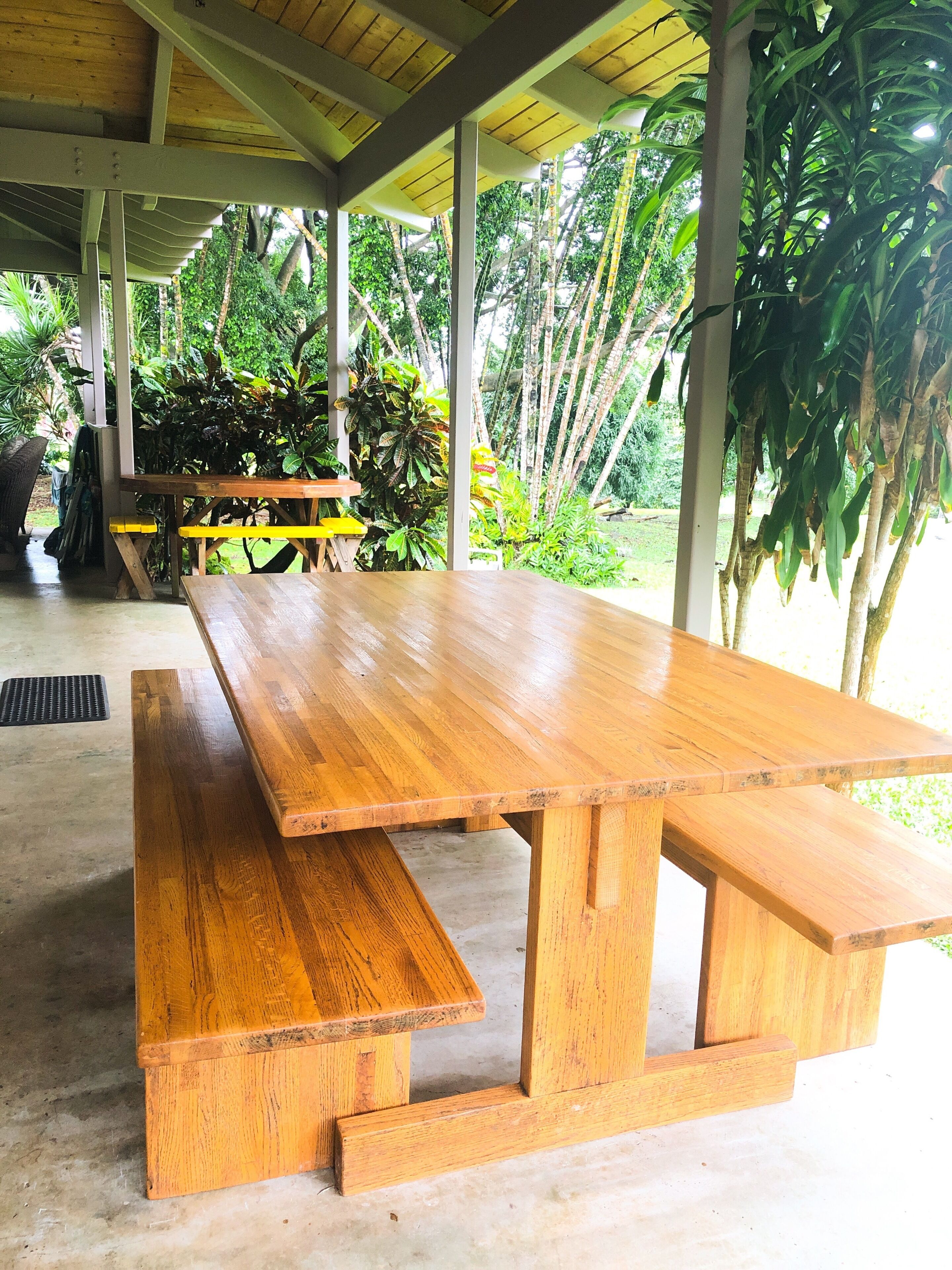 A wooden picnic table with benches under a covered area with a view of tropical vegetation.