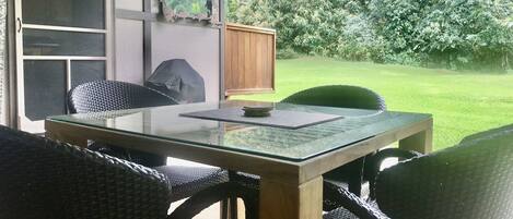 Un patio al aire libre con una mesa de vidrio y sillas de mimbre, una terraza de madera, y vistas a un césped y árboles.