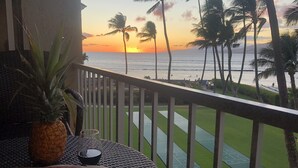 A balcony with a view of the sunset over the ocean, a pineapple, a glass of red wine, and a table with a cutting board and olives.