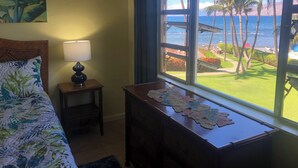 A bedroom with a bed, a bedside table with a lamp, a dresser, and a window with a view of palm trees and the ocean.