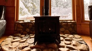 A cozy cabin interior with a stone fireplace, wooden walls, and large windows offering a view of snow-covered trees. - Riverfront, with Hot Tub & Deck Overlooking the River! Sandy beach steps away! (Leavenworth)