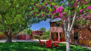 A backyard with a red building, a tree with pink flowers, and red chairs. - The Main House (Cali Cochitta) (Moab)