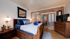A bedroom with a wooden bed, a dresser, a TV, and a brick wall. - The Main House (Cali Cochitta) (Moab)