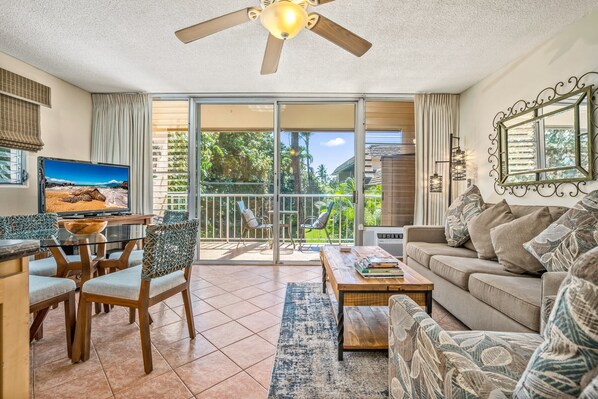 A spacious living room with a large sliding glass door leading to a balcony, a ceiling fan, a television, and a sofa with patterned cushions.