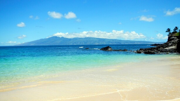 A beach with clear turquoise water, a sandy shore, and a mountain in the background.