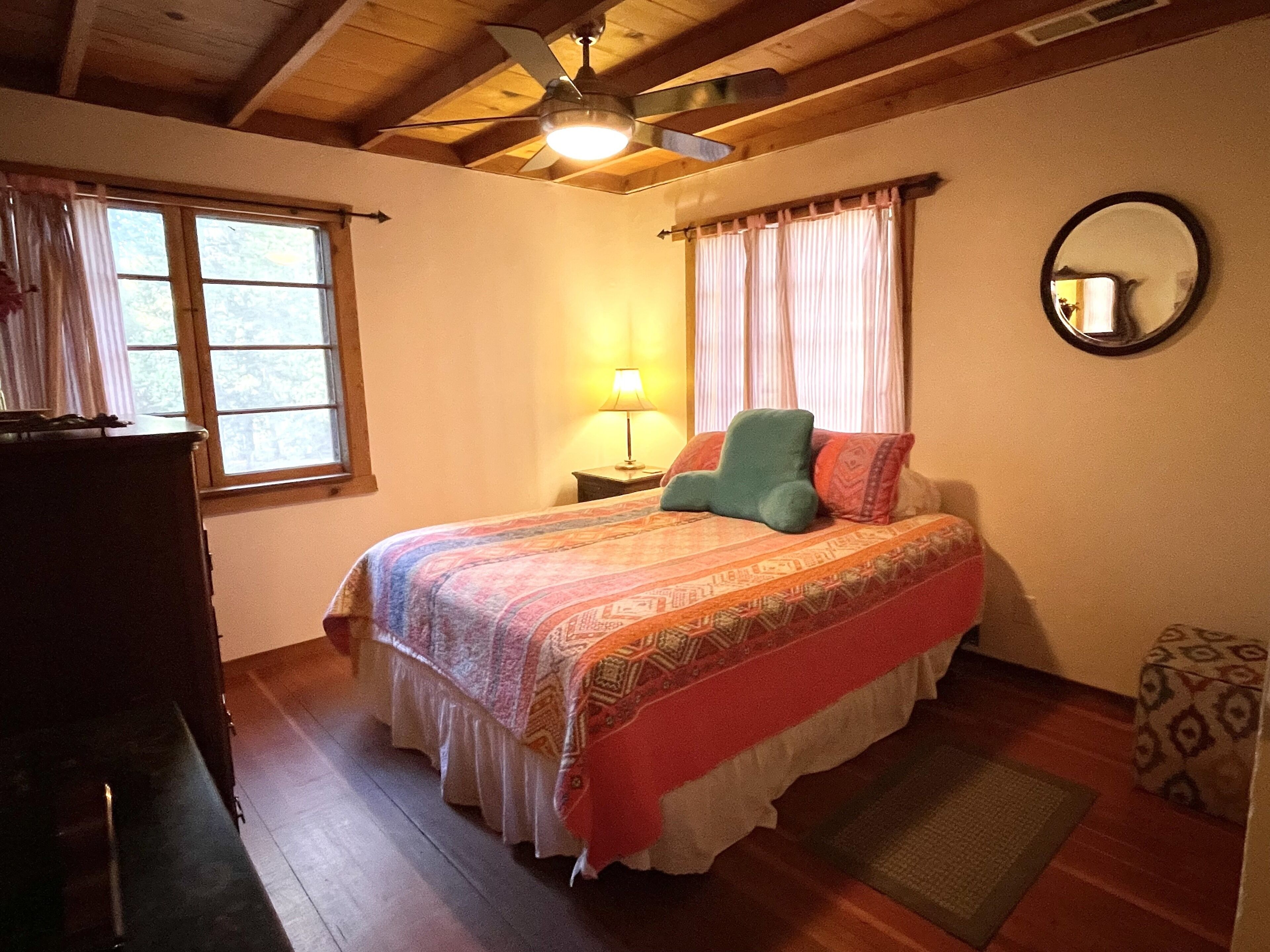 Une chambre à coucher avec un lit, un ventilateur de plafond, une fenêtre avec des rideaux, une table de chevet avec une lampe et un miroir rond accroché au mur.