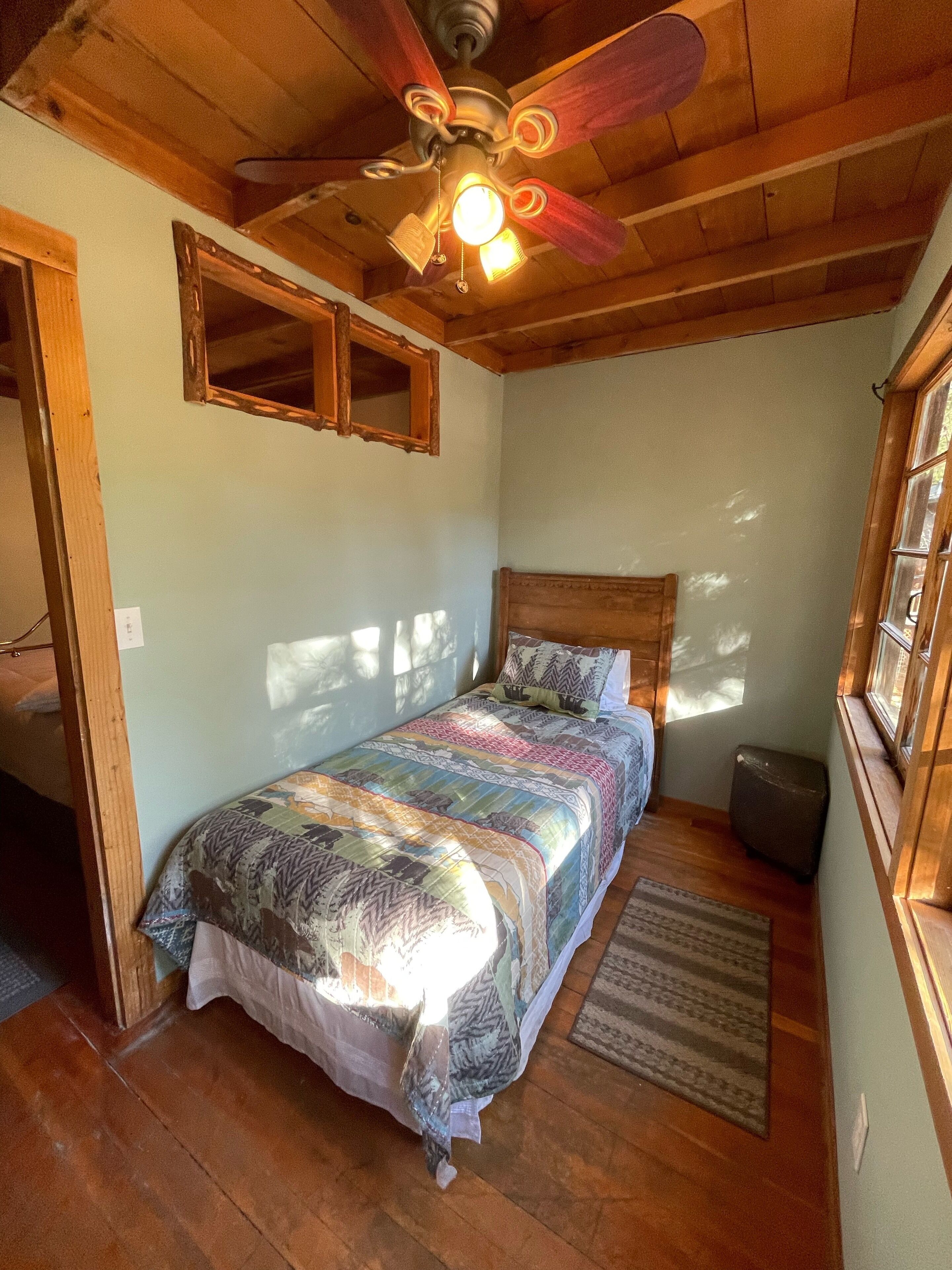 Une chambre avec un plafond en bois, un lit recouvert d’une couvre-lit à motifs, une fenêtre et un ventilateur de plafond.
