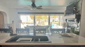 A kitchen with a large window, a ceiling fan, a sink, and a dining area with chairs and a table.