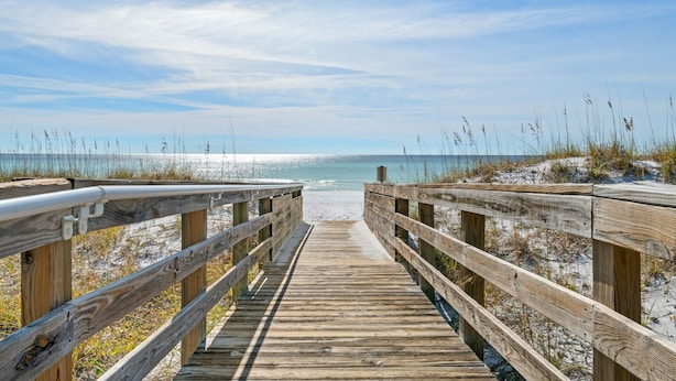 Une promenade en bois qui mène à une plage bordée de dunes de sable et d’une végétation clairsemée.
