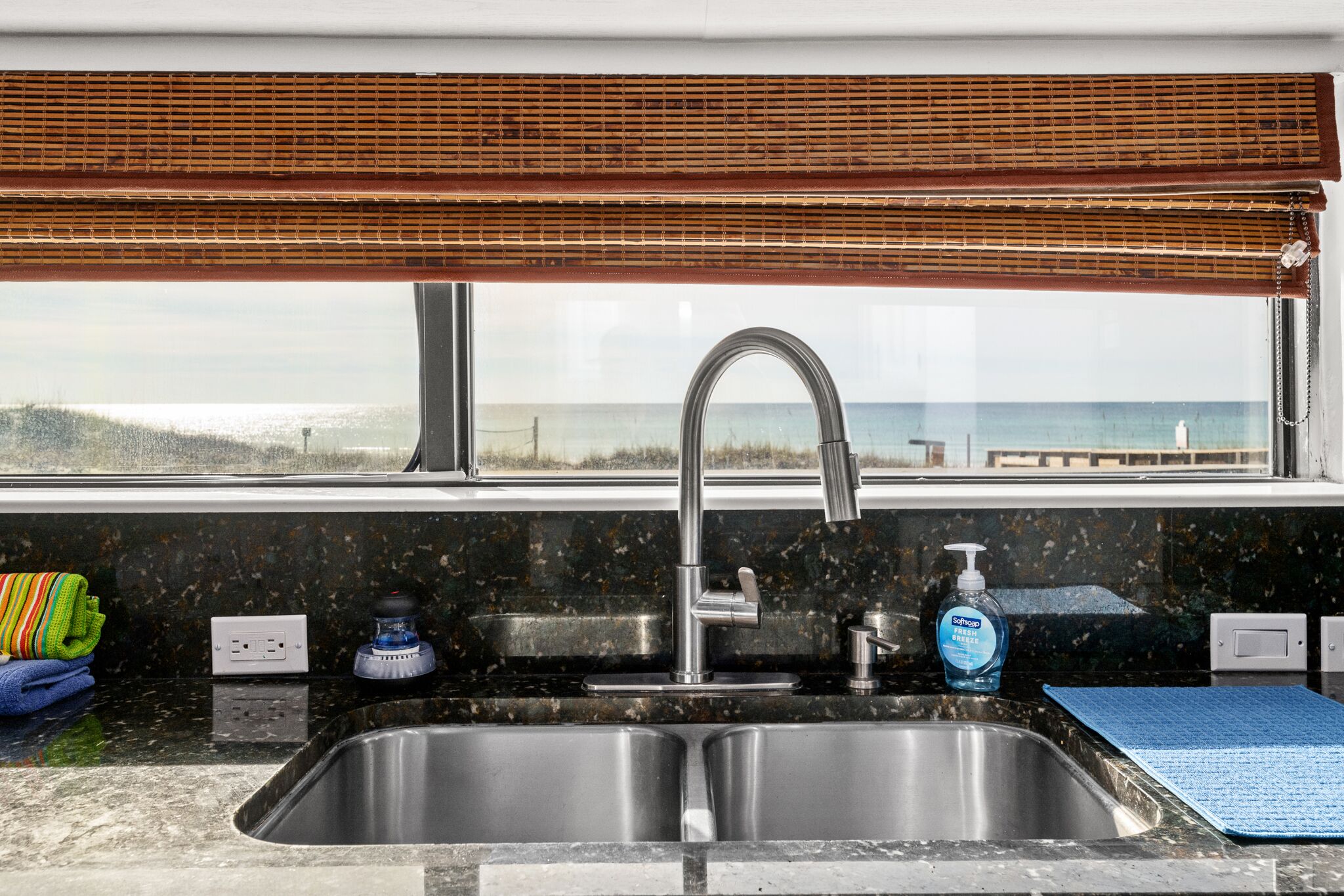 A kitchen with a stainless steel sink, a window with a view of the beach, and a bottle of soap on the counter.