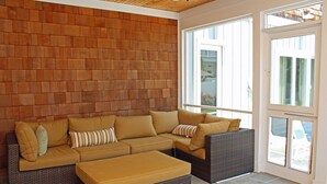 Ein gemütlicher Wohnbereich mit einem Ecksofa, einem Hocker, einer Holzwand und großen Fenstern.