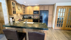 A kitchen with wooden cabinets, a stainless steel refrigerator, a microwave, a toaster, and a sink. There are two bar stools and a clock on the wall.