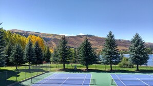 Een tennisbaan met net, omringd door bomen en een meer, met bergen op de achtergrond.