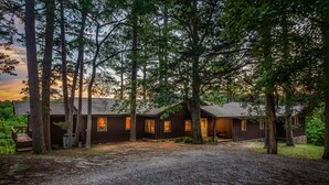 Un chalet rustique avec une allée en gravier, entouré de grands arbres et illuminé par un coucher de soleil en arrière-plan.