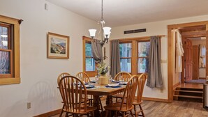 Une salle à manger avec une table en bois dressée pour quatre personnes, entourée de chaises en bois. Un lustre est suspendu au-dessus de la table, et une fenêtre avec des rideaux se trouve à gauche. Une inscription au-dessus de la fenêtre porte le texte suivant : « Simples bienfaits ».