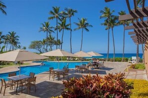 Ein Poolbereich mit Liegestühlen und Sonnenschirmen, umgeben von Palmen und freiem Blick auf das Meer.