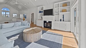 Ein geräumiges Wohnzimmer mit einer großen Couch, einem runden Couchtisch und einem Kamin. Über dem Kamin ist ein Flachbildfernseher angebracht und eine Bücherwand mit diversen Gegenständen.