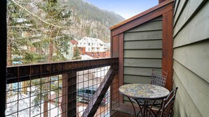 Een balkon met een metalen hekwerk, een tafeltje en stoelen. Er is uitzicht op een besneeerd landschap en gebouwen op de achtergrond.